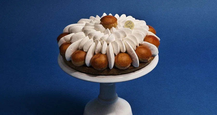 Dalloyau Saint-Honoré mit karamellisierten Windbeuteln und gespritzter Creme auf einer weißen Etagere vor tiefblauem Hintergrund